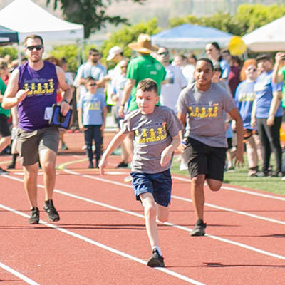 A group of people, some wearing matching t-shirts, are running on a track surrounded by tents and spectators in a park-like setting.