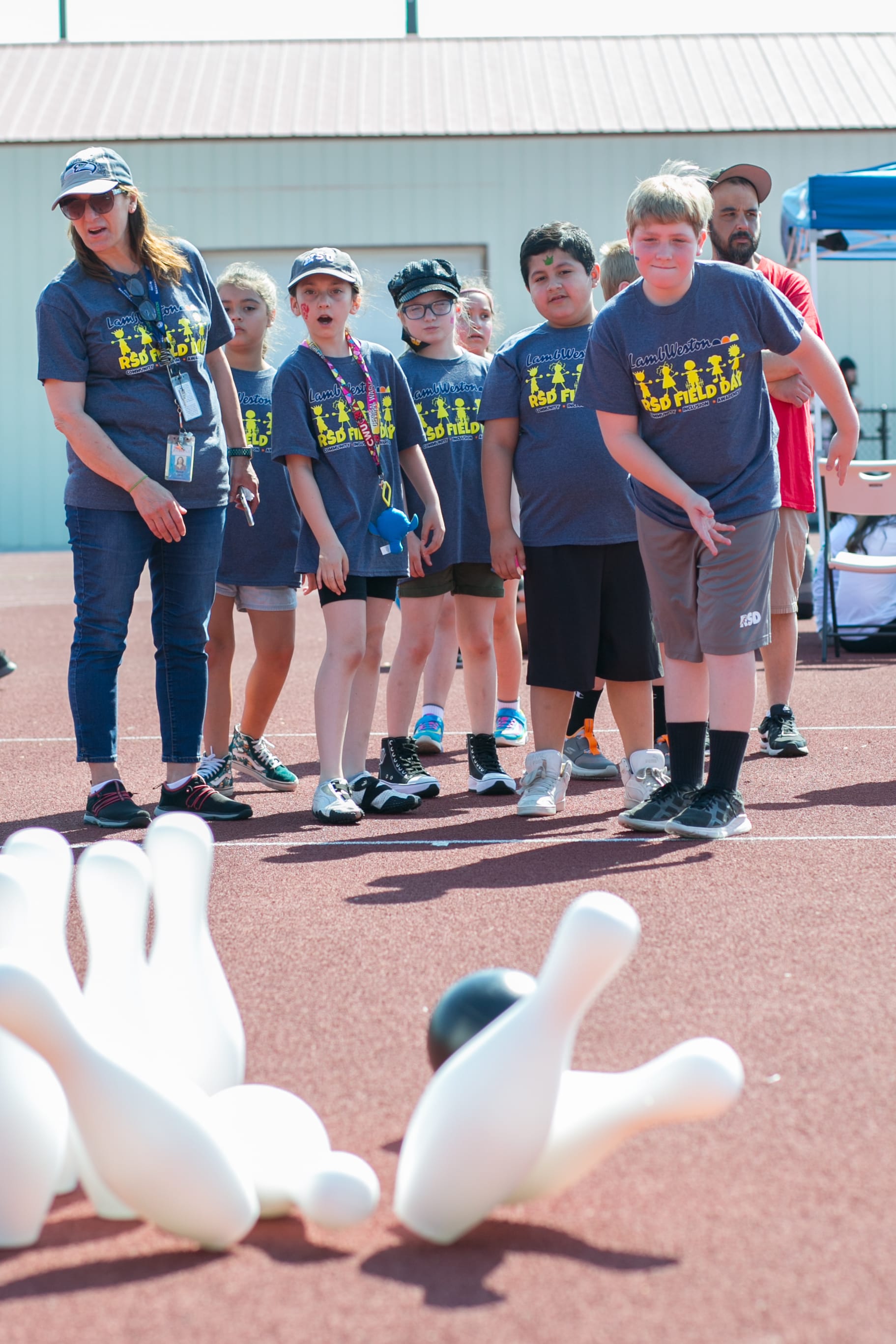 A group of young children wearing matching t-shirts with a crown logo stand on a sports court, with large inflatable bowling pins in the foreground.