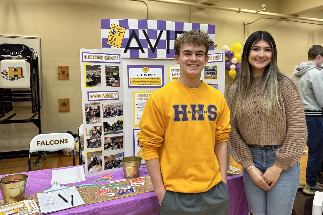 Two young people, a man and a woman, are standing in front of a display board with various school-related items and information.