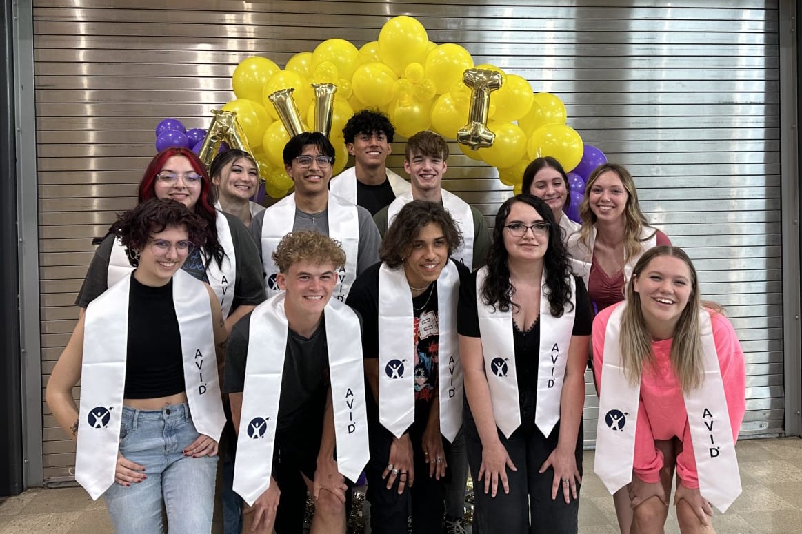 A group of young people, some wearing black and white robes, stand in front of a backdrop of yellow balloons, appearing to celebrate or commemorate an event.