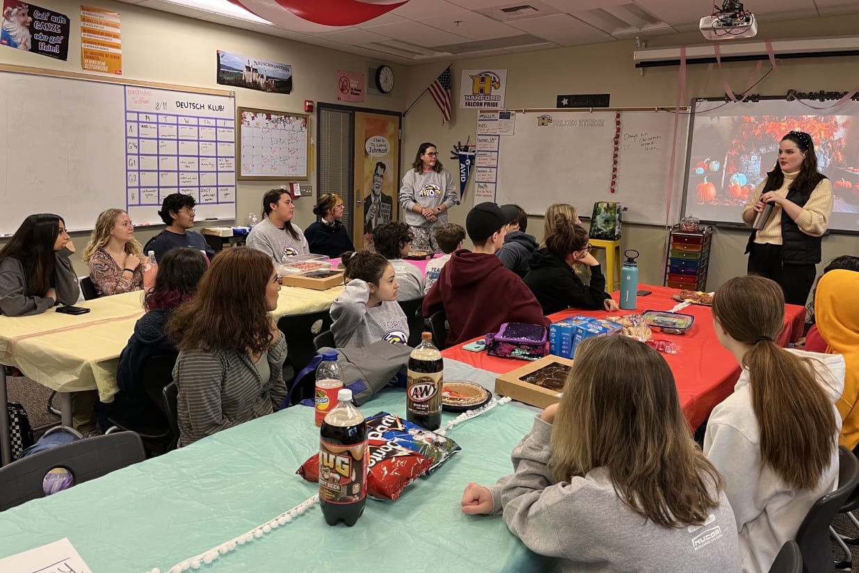 A classroom setting with students gathered around tables, engaged in various activities and discussions, with the teacher standing in the front of the room.