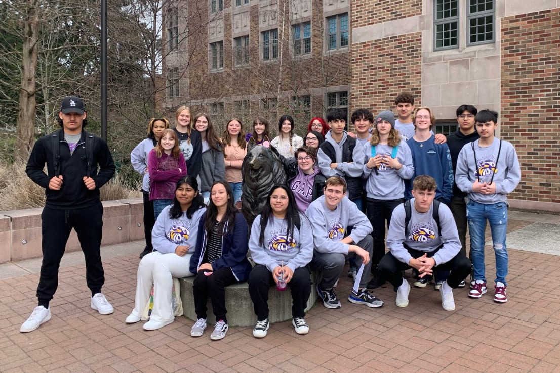 A group of young people, mostly women, are gathered together in front of a brick building, some wearing matching sweatshirts and others in casual attire, creating a sense of community and camaraderie.