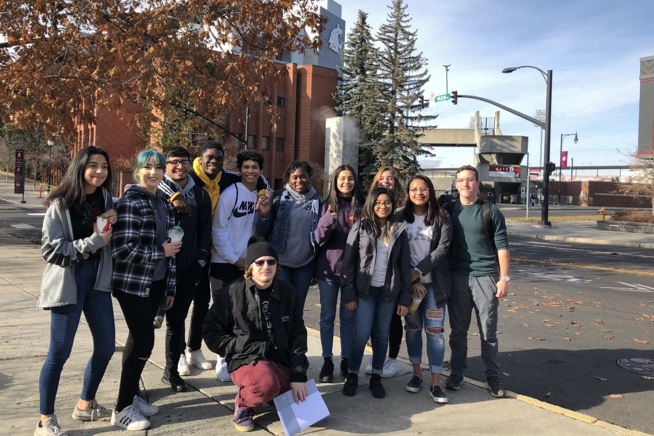 A group of young people, mostly women, standing together outdoors in an urban setting with trees and buildings in the background.