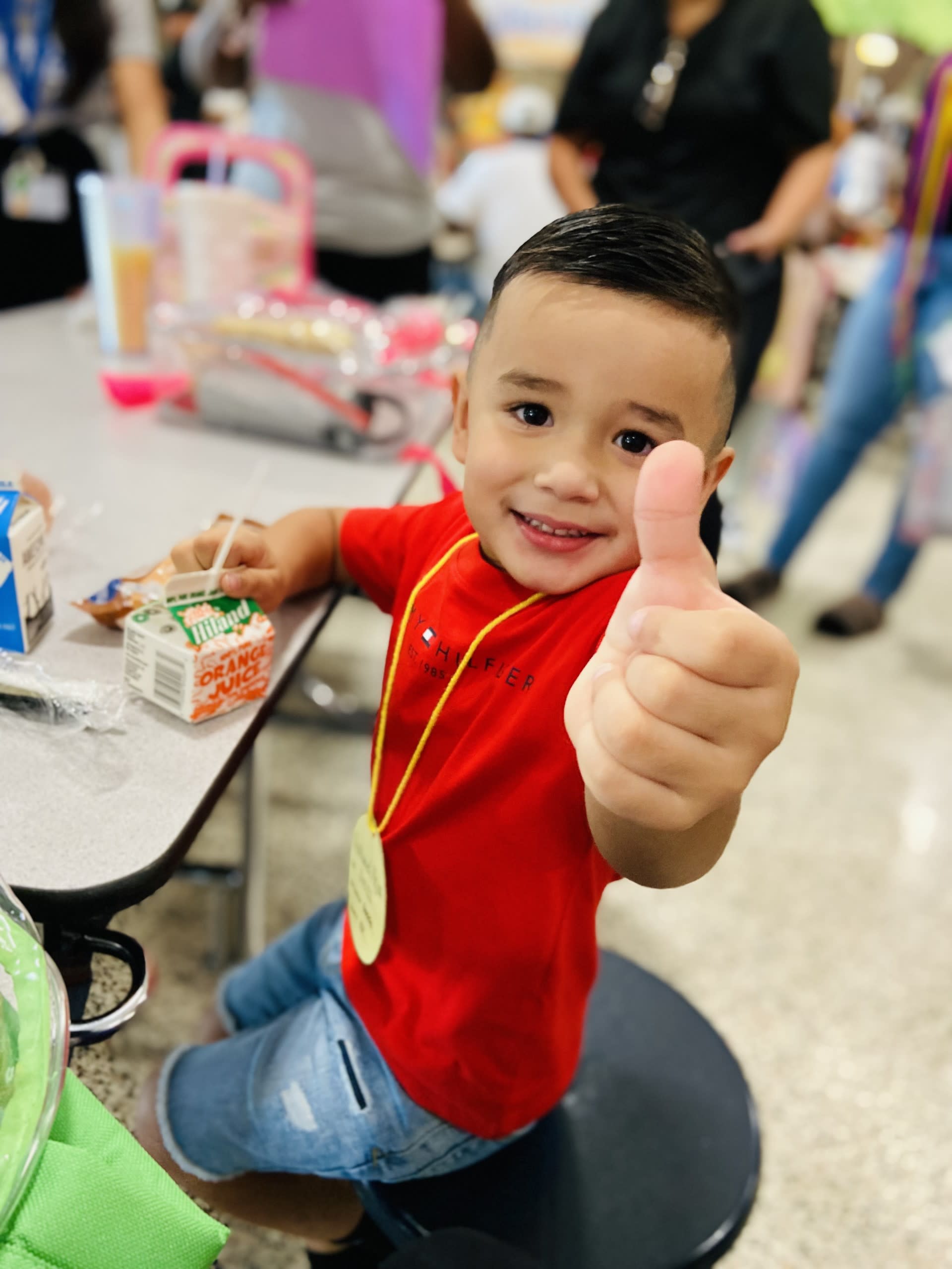 boy in a red shirt seated at cafeteria table giving a thumbs up.