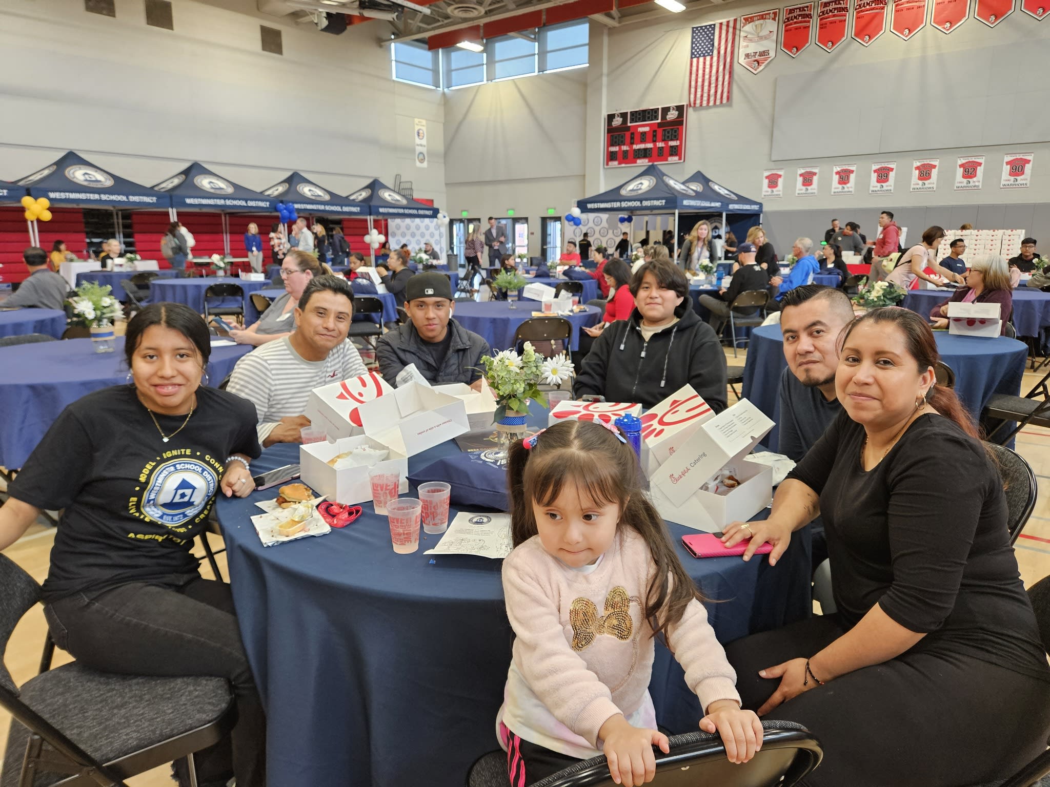 A crowded indoor event with various tents, people sitting at tables, and a young girl in the foreground.