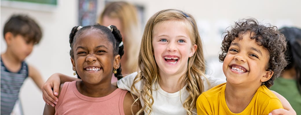 Three smiling children of diverse backgrounds are the focus of the image, which appears to be taken in a classroom or educational setting with colorful artwork and posters visible in the background.