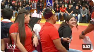 A group of students sit on chairs in a gymnasium during a school event, with spectators watching in the background.