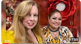 Two women smiling, standing indoors in front of a large red flower decoration on a wooden wall.