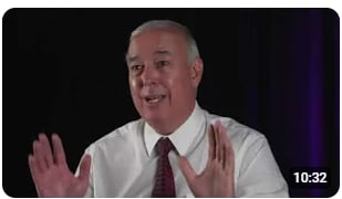 A man in a white shirt and tie is speaking, gesturing with his hands to emphasize his points.