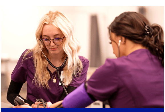 Two women dressed in purple scrubs are engaged in examining a stethoscope, reflecting their roles in the medical field.