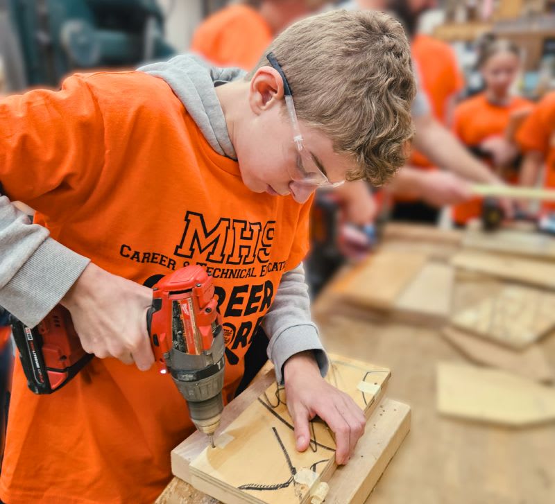 CTE student in an orange MHS shirt using a power drill in a woodworking class.