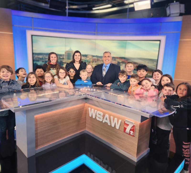 Elementary students visiting WSAW 7 news studio, posing with anchors at the news desk.