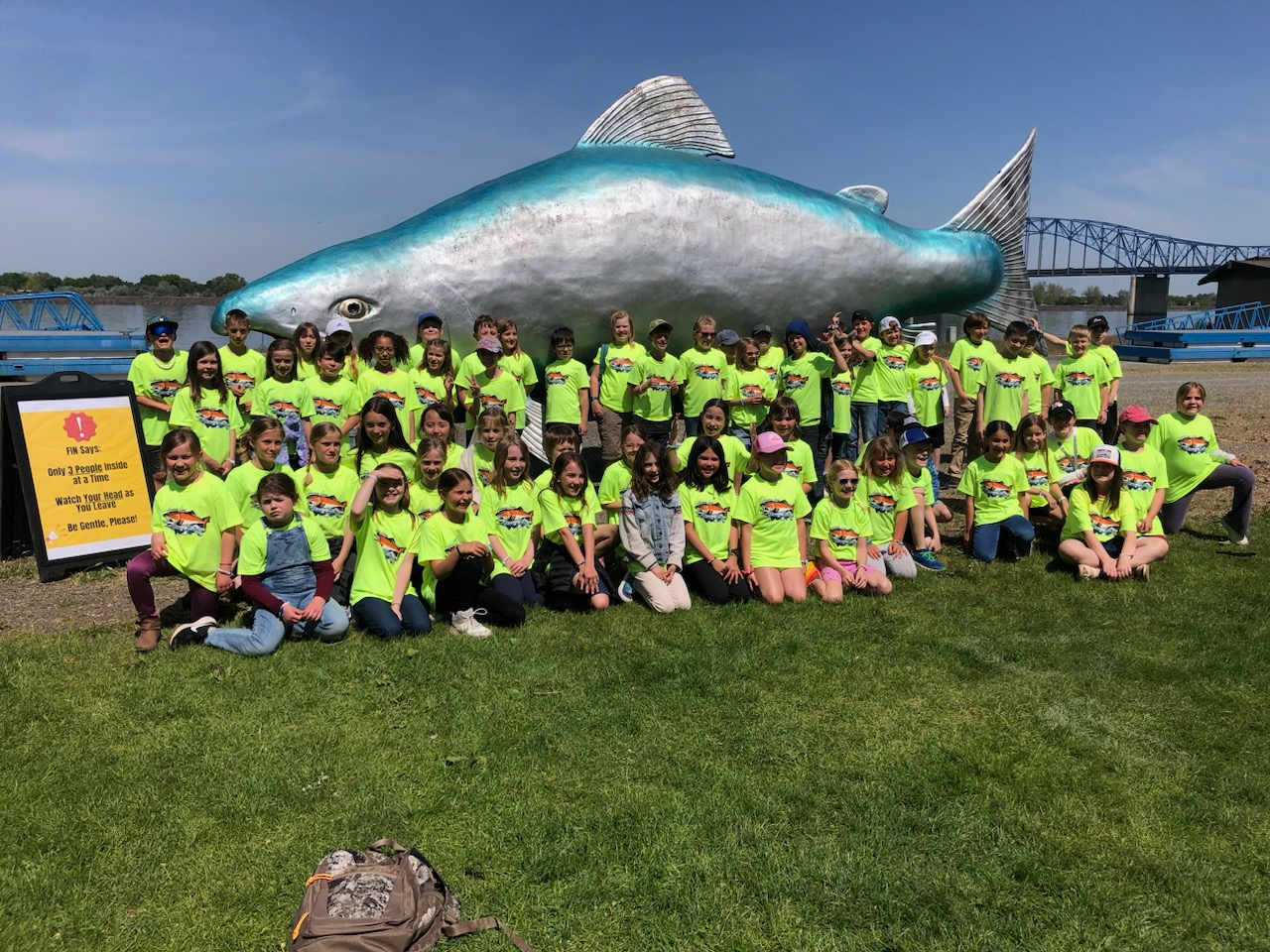 A large group of people wearing bright yellow shirts stand in front of a giant, colorful sculpture of a fish, with a bridge visible in the background.