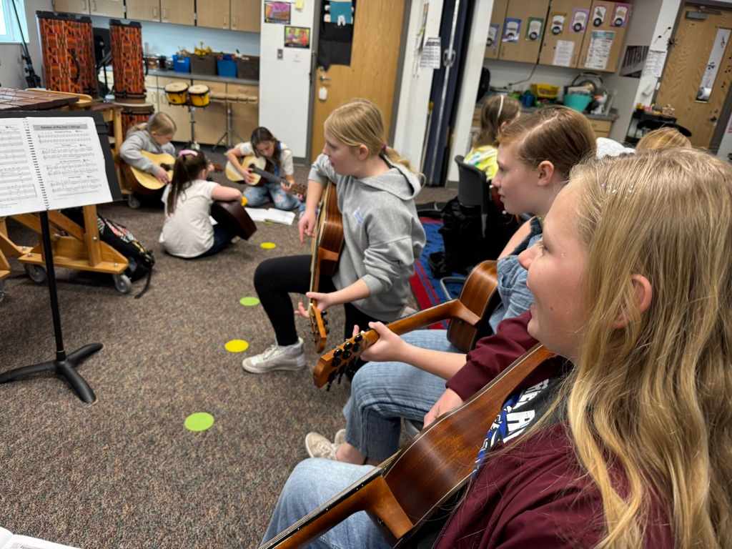 A group of students, some playing guitars, are gathered in a classroom setting with various educational materials and supplies visible in the background.