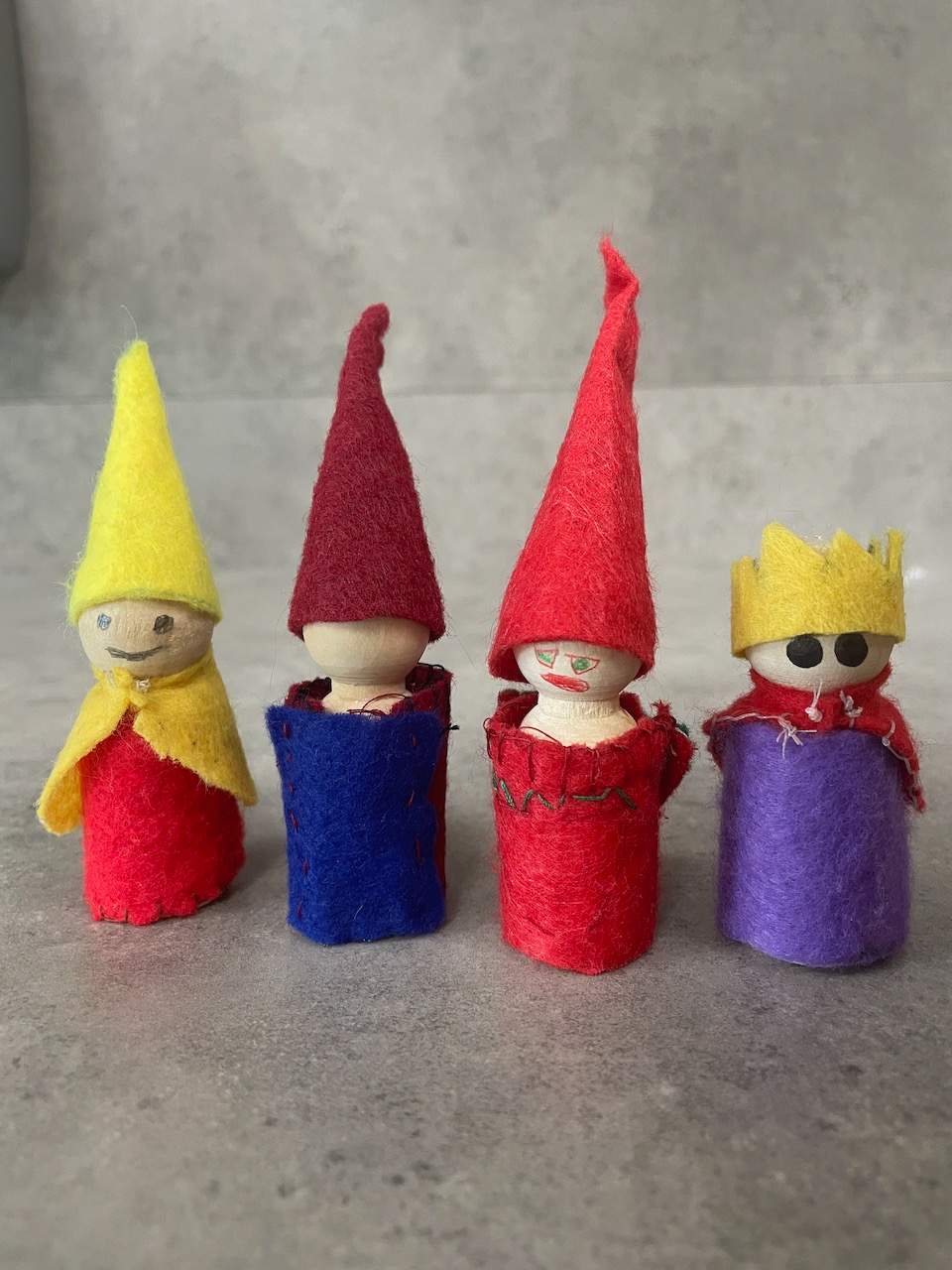 Four colorful felt gnome figurines with pointed hats and smiling faces are displayed on a gray concrete surface.