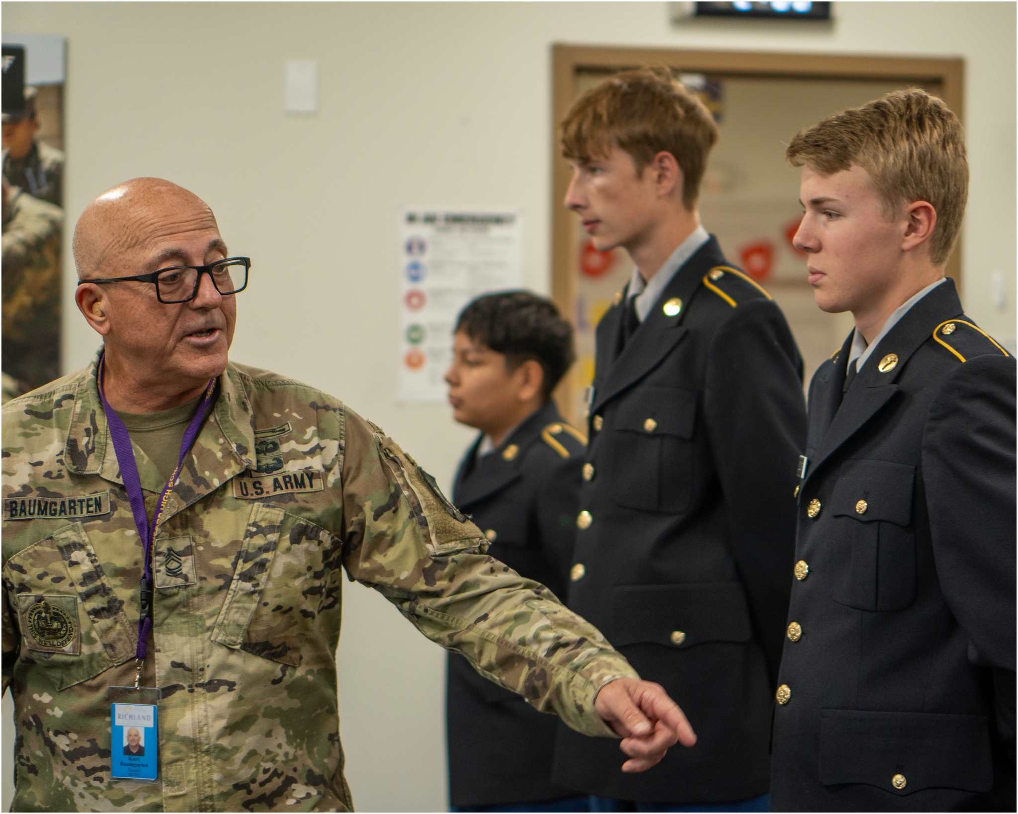 A military officer in camouflage uniform is speaking with three young men in military uniforms in what appears to be an indoor setting with a bulletin board in the background.