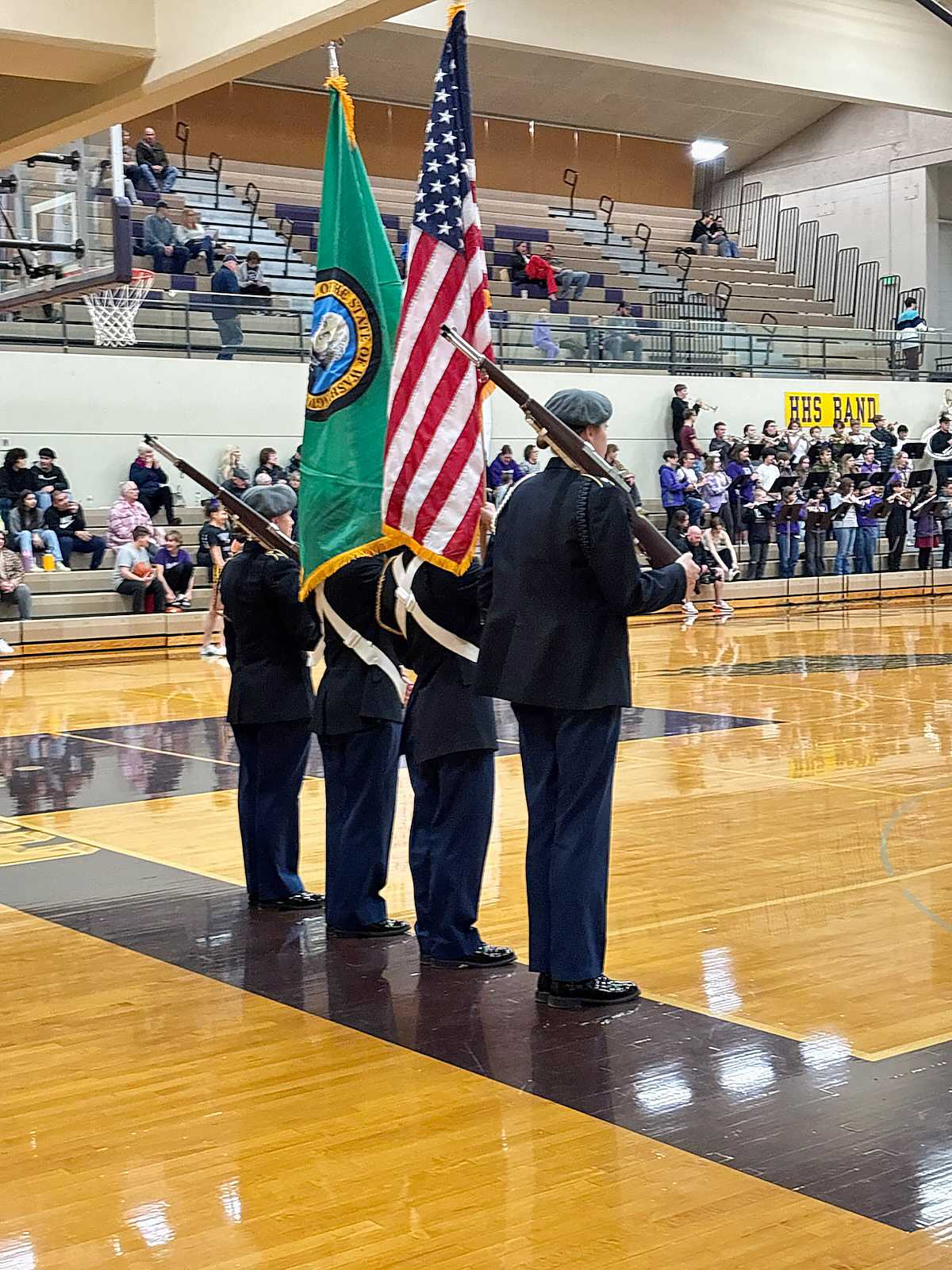 A group of uniformed individuals holding the American and Washington state flags stands on a basketball court in front of a crowd of spectators in a gymnasium.