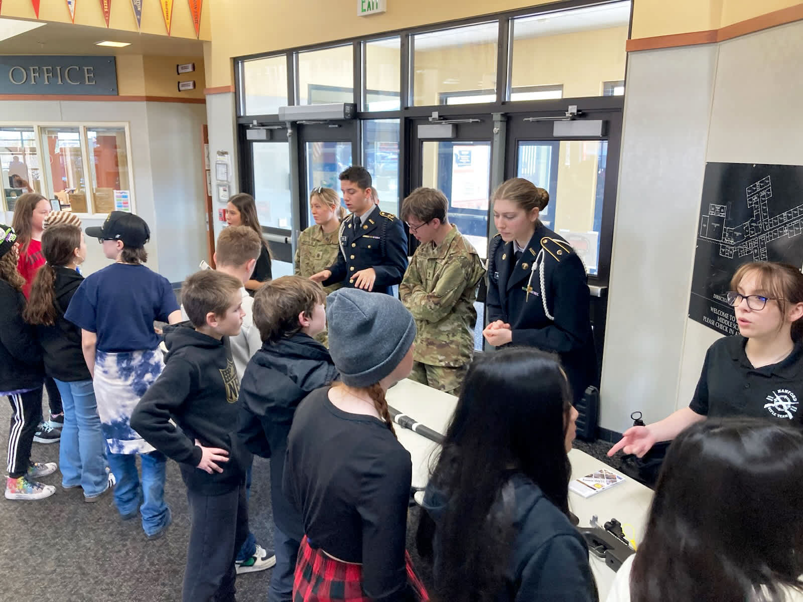 A group of people, mostly young individuals, are gathered in what appears to be a school or community center setting, with various posters and signage visible in the background.