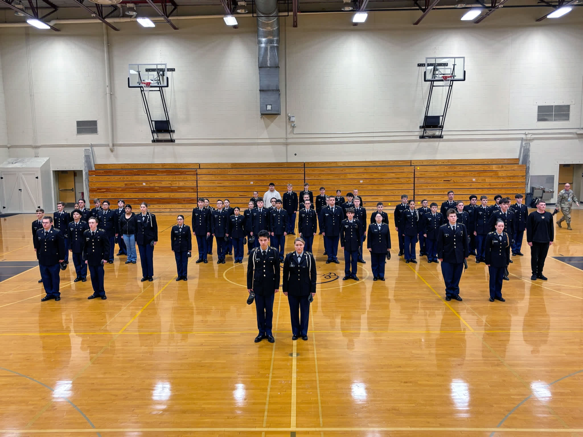 A large group of uniformed individuals, likely members of a military or law enforcement organization, standing in a gymnasium-like indoor setting with basketball hoops visible in the background.