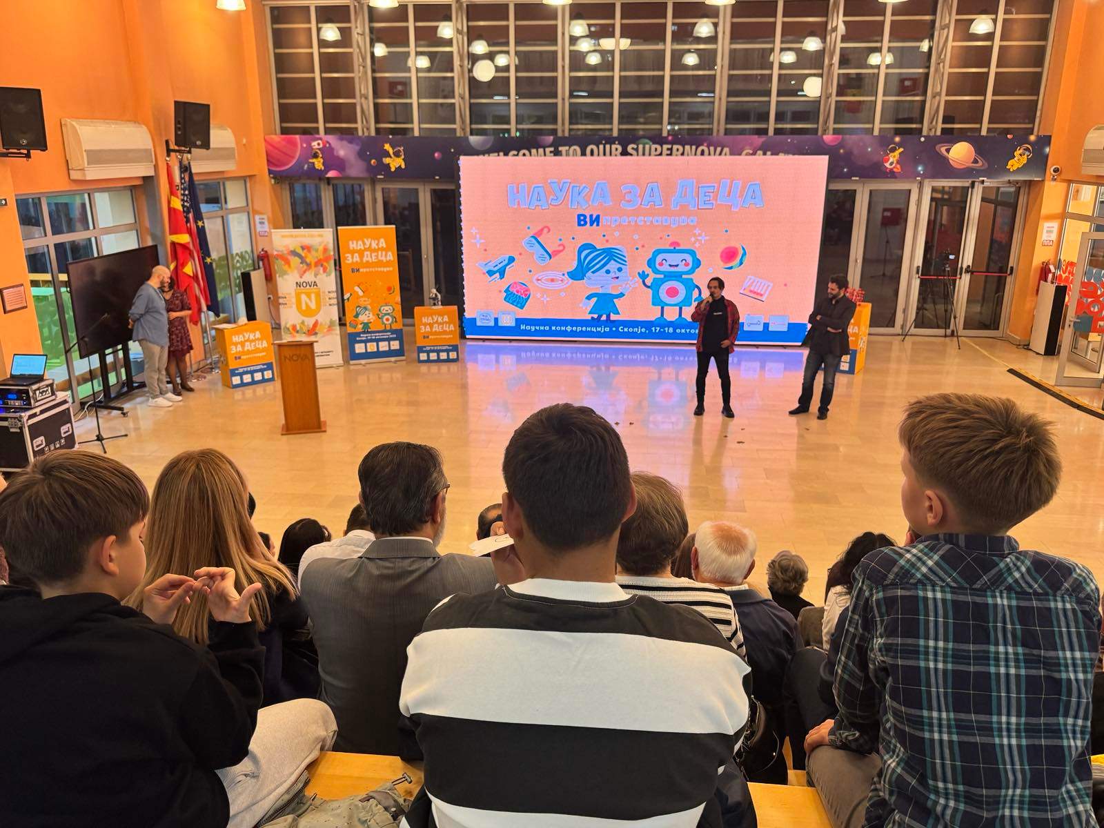 A group of people, mostly young, are gathered in a large room with colorful displays and signage on the walls, suggesting an educational or informative event.