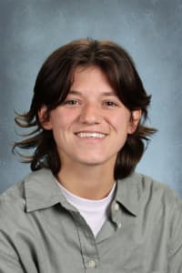 A smiling young person with short, dark hair and a collared shirt is centered against a mottled blue background.