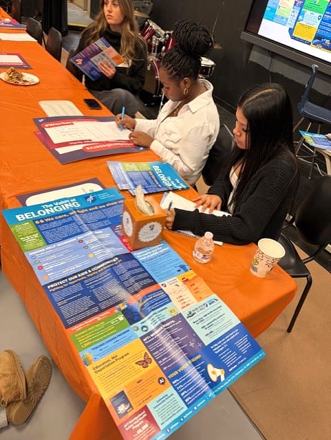 Students seatwed at table with orange cover and with literature on it