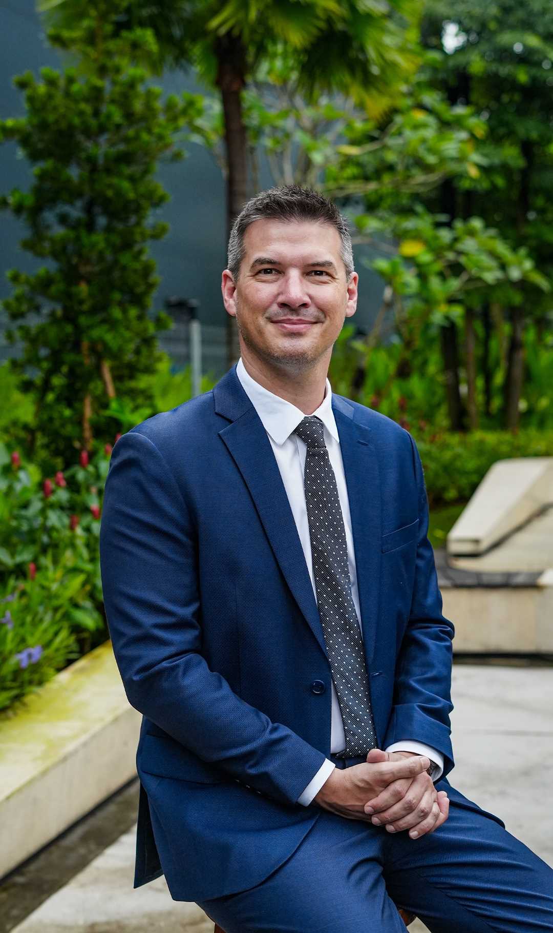 A well-dressed man in a navy blue suit sitting on a bench in a lush green outdoor setting