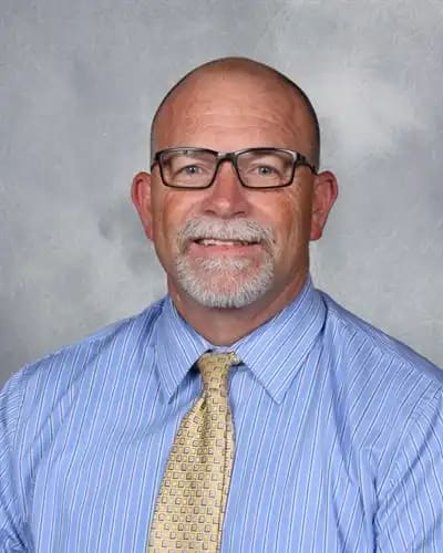 A smiling man with glasses and a blue striped shirt with a yellow tie is in the foreground, against a gray, mottled background.