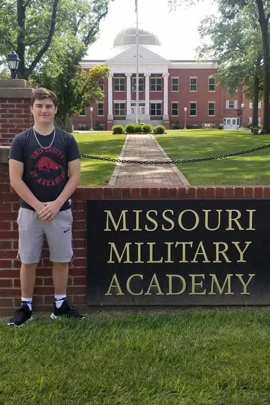 Young man stands in front of Missouri Military Academy sign with campus building in background.