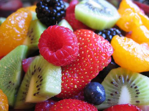 A vibrant close-up of a fruit salad features a bright red raspberry and strawberry in the foreground, with slices of kiwi, orange segments, blackberries, and blueberries filling the background.