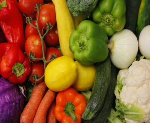 A vibrant assortment of fresh vegetables, including tomatoes, peppers, lemons, and carrots, fills the foreground, while broccoli and cauliflower are visible in the background.