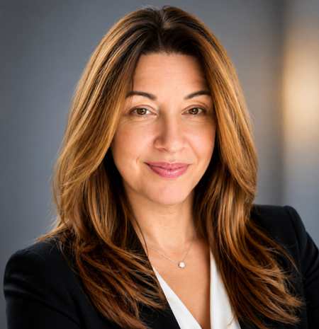 A woman with shoulder-length brown hair and a black blazer smiles at the camera against a blurred gray background.