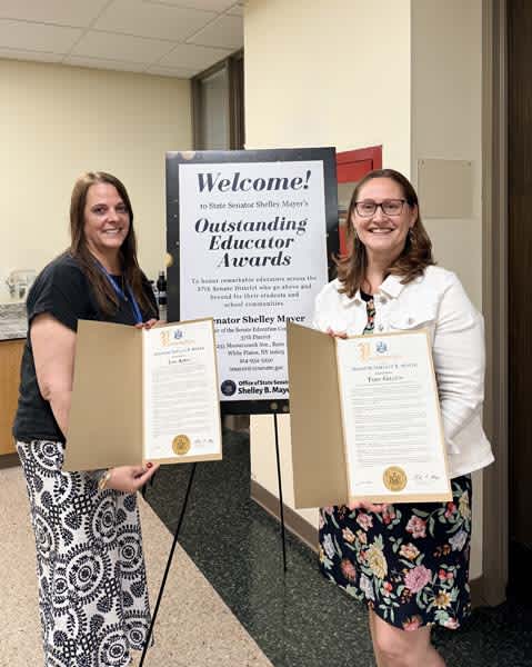 Two women stand in front of a welcome sign for Outstanding Educator Awards, holding framed certificates.