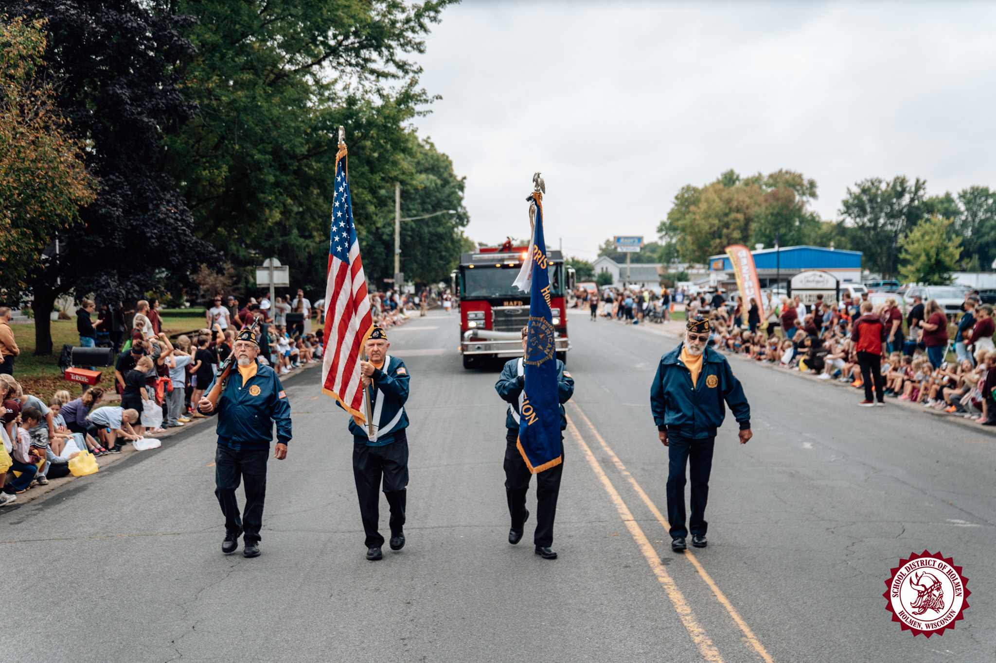 Veterans carry the American flag and a local post flag while leading the Holmen Homecoming Parade through a crowd-lined street.