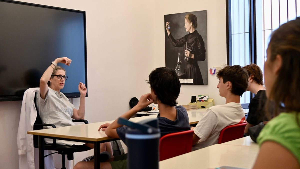 A teacher gestures while speaking to students in a classroom with a portrait of Marie Curie on the wall.