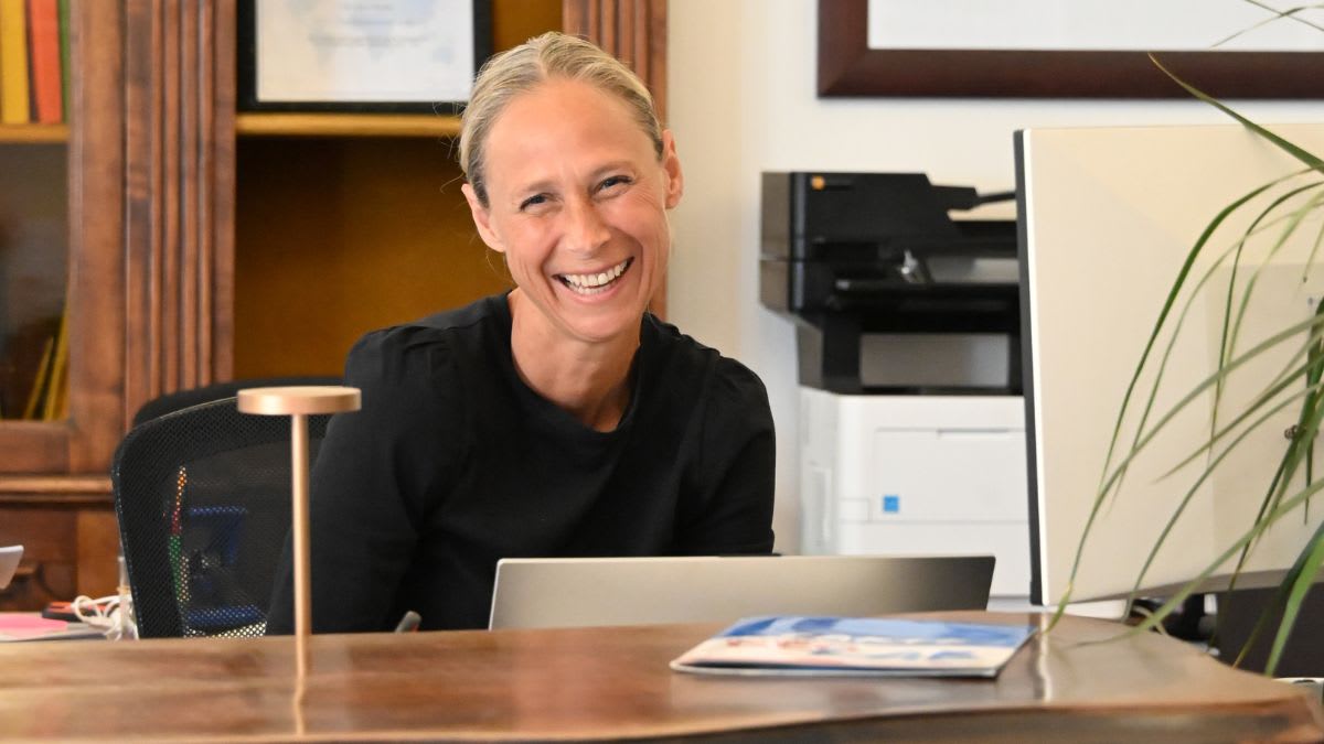A smiling woman sits at a desk with a laptop and a lamp in the foreground, while a printer and bookshelves are visible in the background.