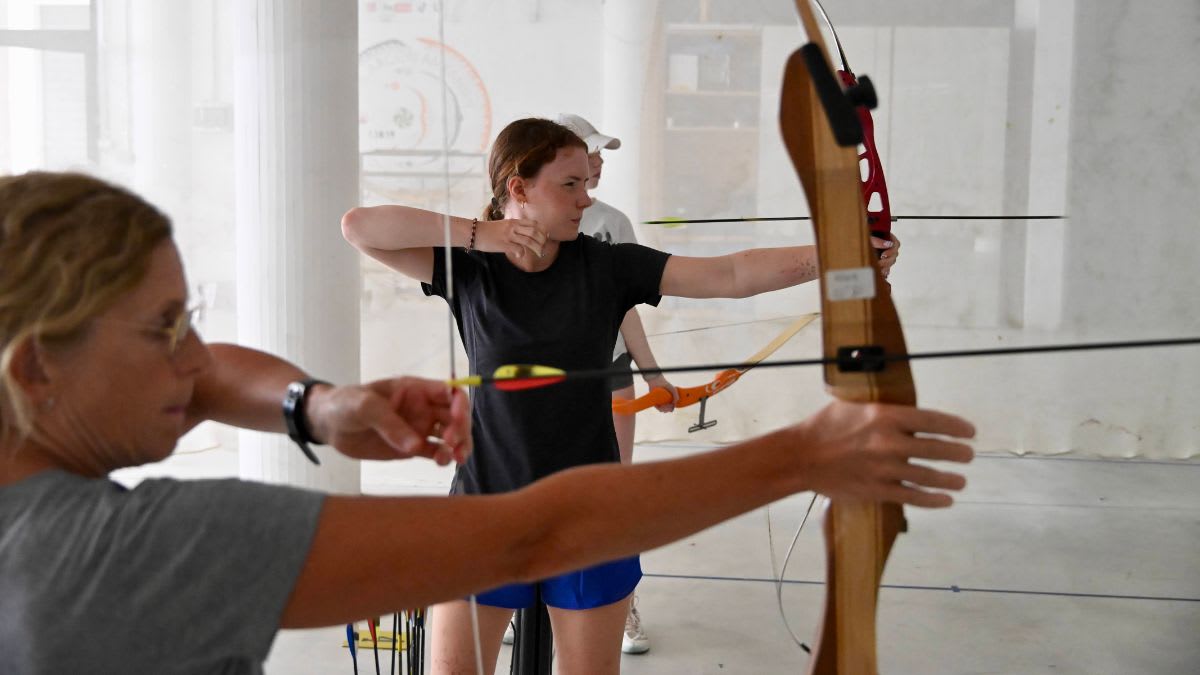 In the foreground, a woman with blonde curly hair draws back a bowstring, while in the background, another woman in a black shirt aims her arrow.