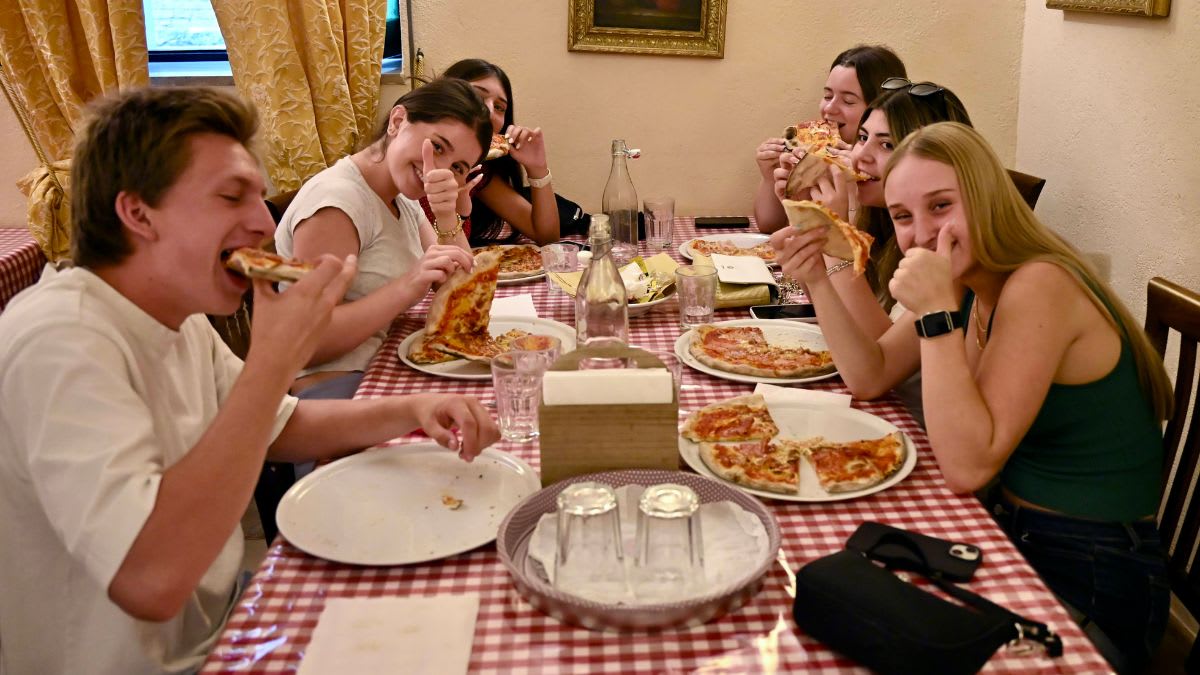 A group of friends enjoys pizza at a restaurant with a checkered tablecloth in the foreground and ornate curtains and a painting in the background.