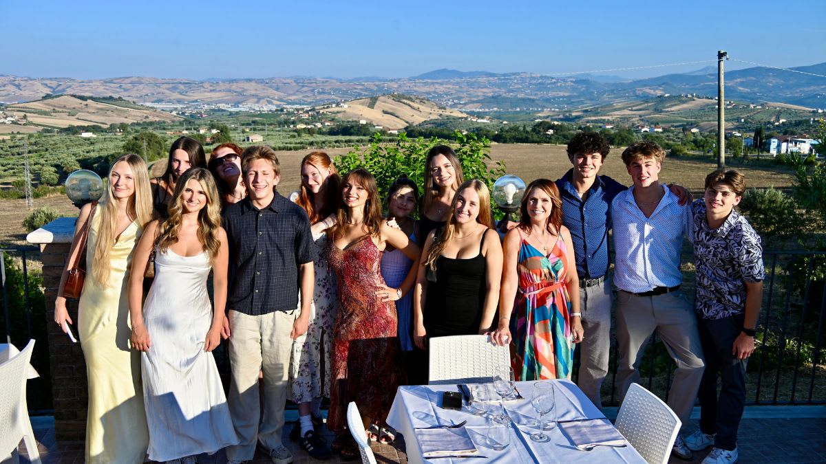 A group of young people in formal attire pose for a photo in the foreground, with a scenic, rolling landscape of hills and scattered buildings in the background.