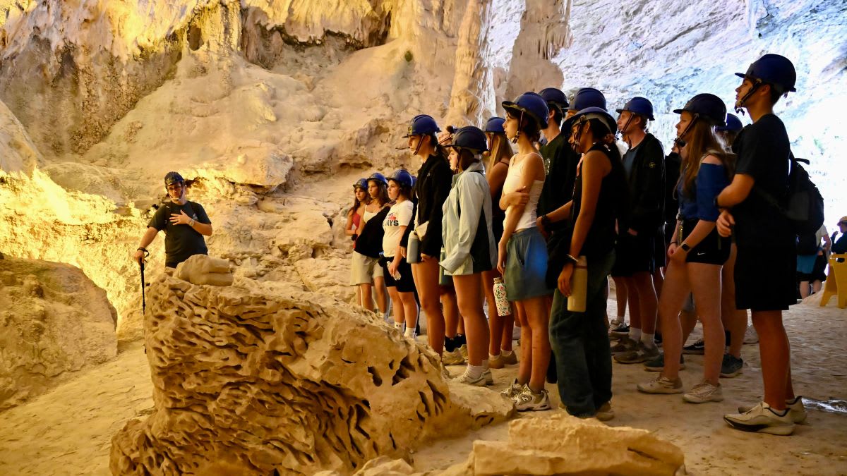 A tour guide in a cave gestures to a group of tourists wearing hard hats, with the cave's textured walls and ceiling visible in the background.