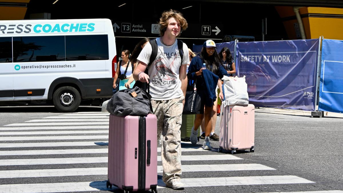 A young man with a backpack and pink suitcase crosses a crosswalk in front of a white bus and a building with a yellow facade.