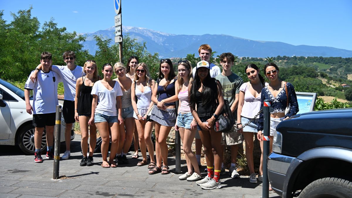 A group of young people stands on a paved area in the foreground, with a scenic mountain range and lush green hills in the background under a clear blue sky.