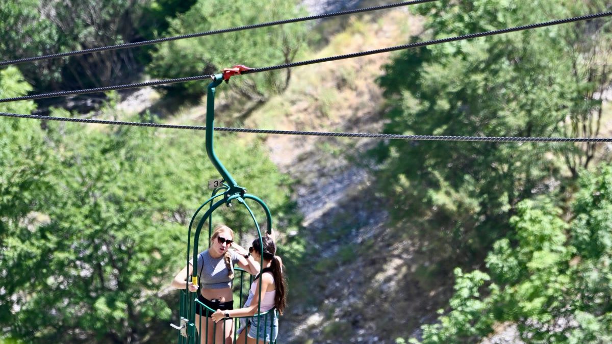 Two young women ride a ski lift through a lush green forest.