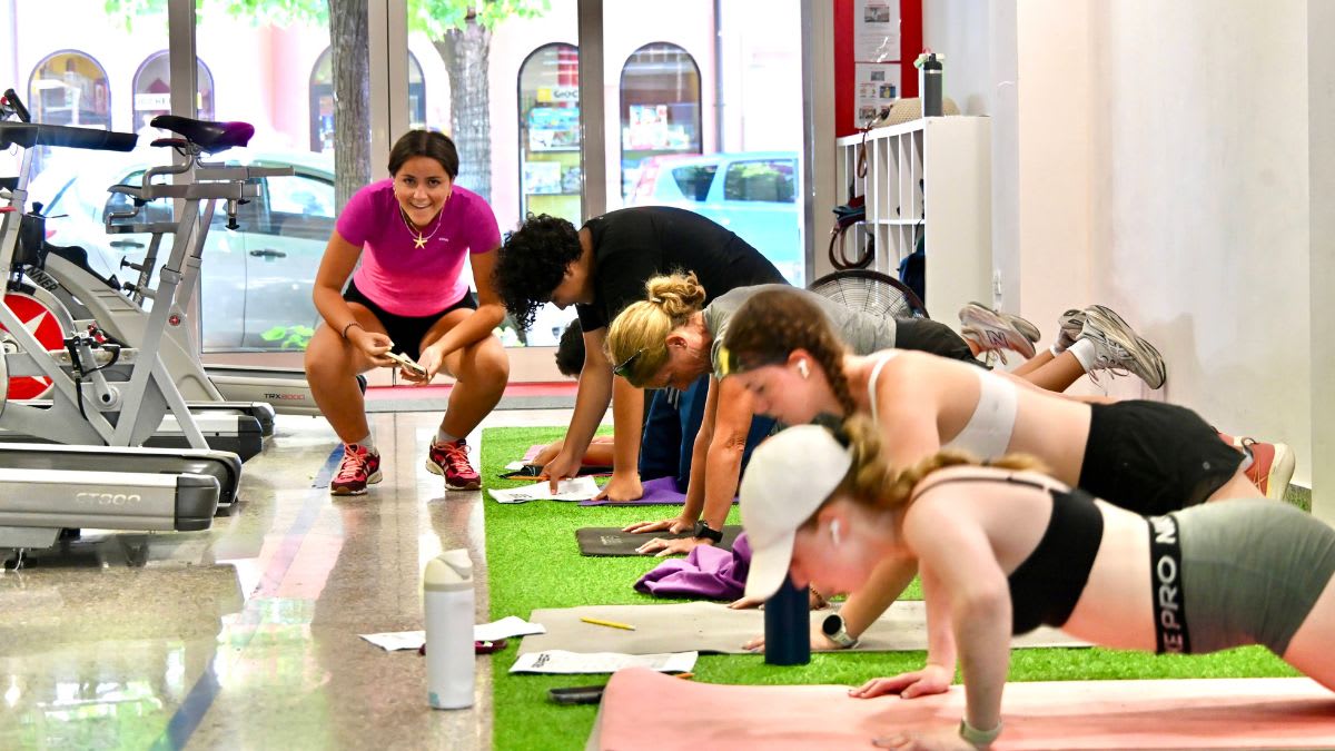 A group of young women perform push-ups on a green mat in the foreground, while a trainer watches and stationary bikes are visible in the background.