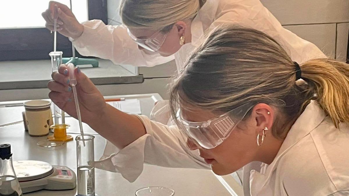Two young women in lab coats and goggles are performing an experiment with beakers and droppers in a laboratory setting.