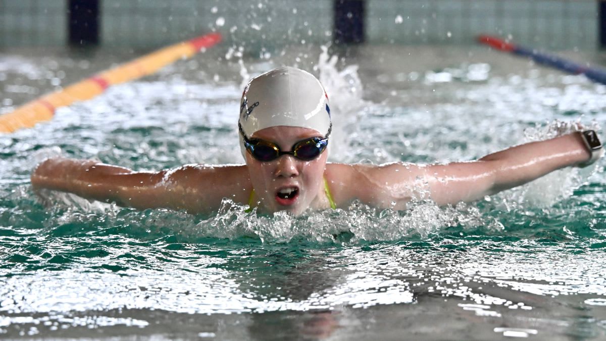 A young swimmer in a white cap and goggles performs the butterfly stroke in a pool, with lane markers visible in the blurred background.