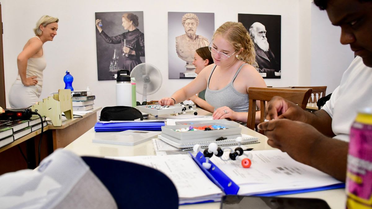 In the foreground, students are engaged in a molecular modeling activity at a table, while in the background, portraits of historical figures adorn the wall.