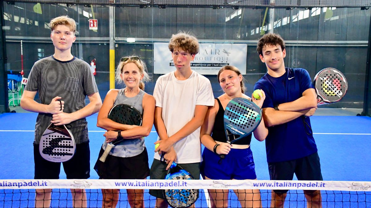 Five people, four holding padel rackets and one holding a tennis ball, stand on a blue padel court with a net in the foreground and a banner in the background.