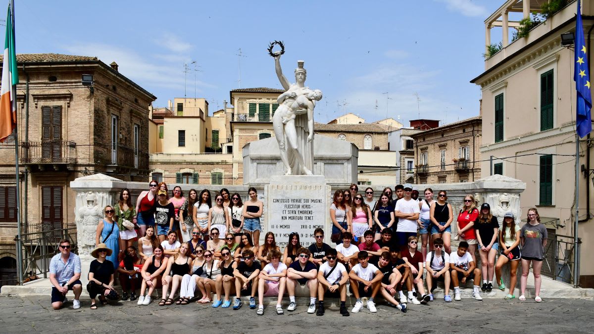 A large group of young people poses for a photo in front of a white monument with a statue, set against a backdrop of Italian buildings under a bright sky.
