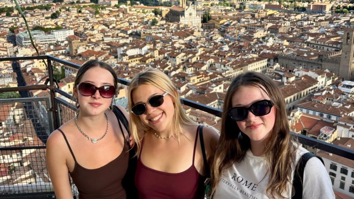 Three young women in sunglasses smile at the camera in the foreground, with a sprawling cityscape of terracotta rooftops and distant buildings in the background.